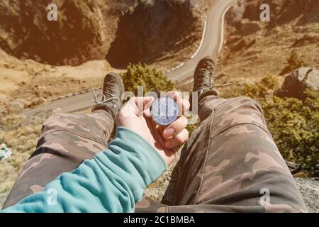 Wanderer auf einem hohen Felsen sitzend hält einen Kompass vor ihren Füßen in Trekkingstiefeln und einer hohen Klippe mit einer asphaltierten Straße darunter. Stockfoto