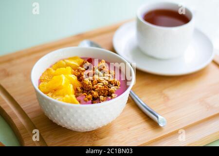 Gesundes Sommerfrühstück einer rosa Smoothie Schale mit gefrorener Banane und Himbeere garniert mit Granola und frischer Orange. Stockfoto