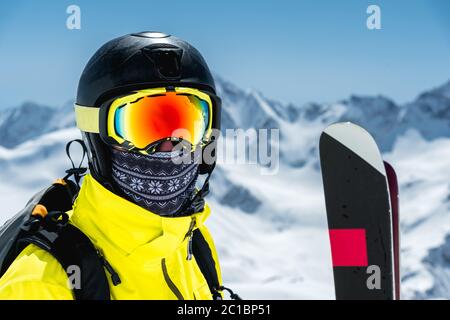 Ein großes Porträt eines Skifahrers in Schutzhelm und Brille - eine Maske und ein Schal neben den Skiern gegen die schneebedeckte mou Stockfoto