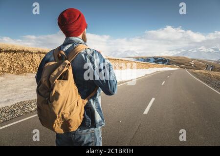 Ein bärtiger Hipster mit einem altmodischen Vintage-Rucksack, der Sonnenbrille mit rotem Hut und Jeans-Jacke und Jeans trägt, steht auf Stockfoto