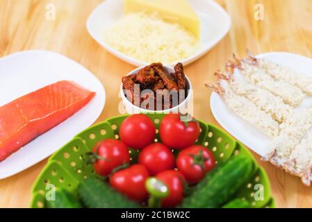 Stillleben roher Lebensmittel in weißen Tellern auf einem Holztisch. Gefrorener Lachs auf einem Teller neben Gurken und Tomaten geriebenen Käse Stockfoto