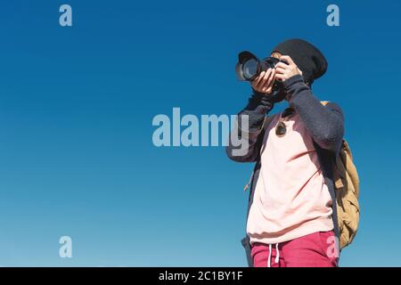 Portrait eines stilvollen Mädchen Hippie in einem Hut und mit einem Rucksack, der Fotos von ihr auf einer DSLR-Kamera im Freien gegen eine blu nimmt Stockfoto