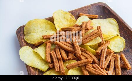 Leckere Kartoffelchips und gesalzene Roggenbrot-Cracker auf einem dunklen Holzteller, auf weißem Hintergrund. Kalorienreicher Snack. Stockfoto