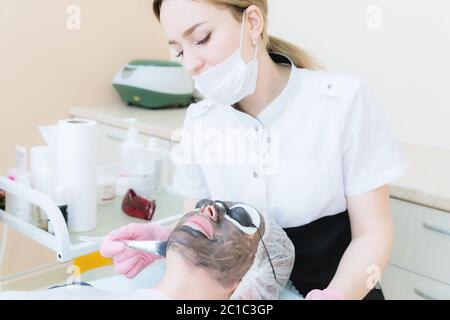 Der Kosmetologe in rosa Handschuhen mit Pinsel trägt eine Carbon-Maske zum Peeling auf das Gesicht eines jungen Mädchens in einem Kosmetologie-Roo Stockfoto