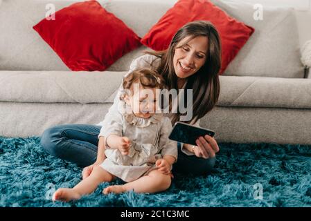 Junge hübsche Mutter spielt mit ihrer Tochter auf dem Wohnzimmerboden mit dem Telefon Stockfoto