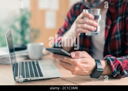 Gig Arbeiter Trinkwasser und mit Handy im Home Office während der freiberuflichen Projektarbeit Stockfoto