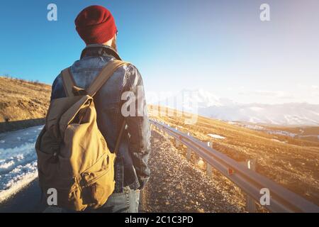 Ein bärtiger Hipster mit einem altmodischen Vintage-Rucksack, der Sonnenbrille mit rotem Hut und Jeans-Jacke und Jeans trägt, steht auf Stockfoto