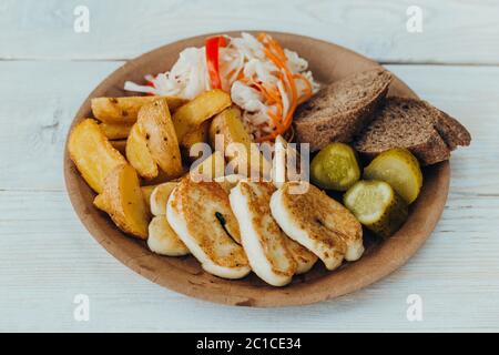 Schmackhafter gegrillter Fisch mit gebratenen Kartoffeln, Gurke, Kohl und Hölzern auf einem Pappteller auf einem Holztisch.Straßenessen Stockfoto