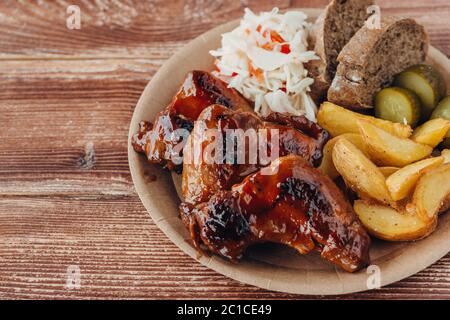Köstliche gebratene Hähnchenflügel mit Sauce, gebratene Kartoffeln, Gurke, Kohl und Khlobs in einem Pappteller auf einem Holztisch. Straßennahrung Stockfoto