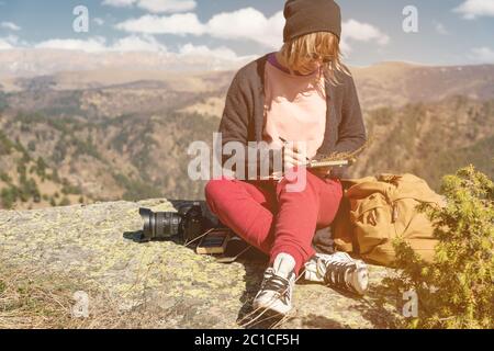 Portrait eines Hipster Mädchens mit Sonnenbrille und Hut auf einem Felsen draußen in den Bergen vor einem blauen Himmel sitzen. Freiberuflich Stockfoto