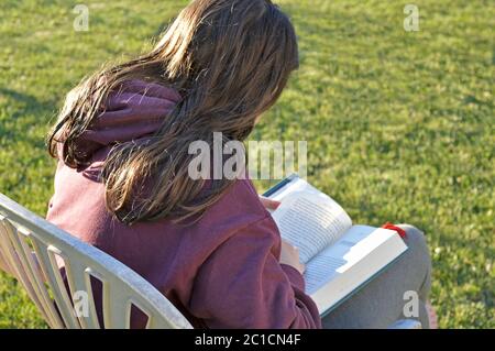 Junge weiße Frau, die in der Sonne sitzt und ein Buch liest. Nicht mit Technologie verbunden Stockfoto