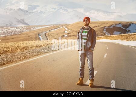 Ein stilvoller bärtiger Hipster in Sonnenbrille mit Vintage-Rucksack steht an einem sonnigen Tag auf einer Landstraße Asphalt. Das Konzept von Stockfoto