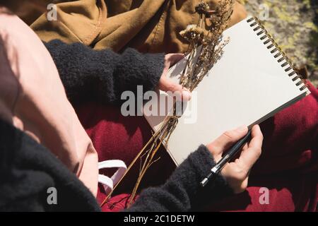 Nahaufnahme der Hände eines Mädchens mit einem leeren Notizbuch. Ein trockener Strauß Kräuter in der Hand und ein Bleistift. Reisedesigner artis Stockfoto