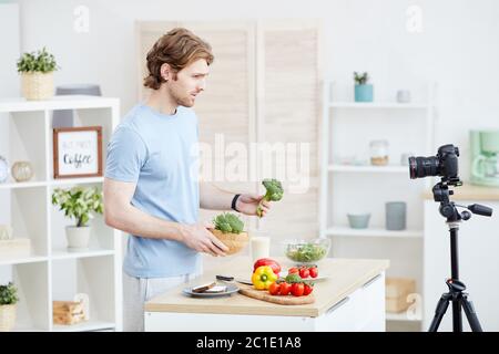 Junger Mann hält Gemüse in der Hand und spricht über die gesunde Ernährung auf der Kamera für seinen Blog Stockfoto