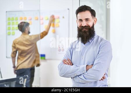 Glückliches Lächeln bärtige Senior Developer oder Manager in modernen IT-Büro. Ein weiterer Profi, der mit Scrum Desk im Hintergrund arbeitet. Erfolgreich Stockfoto