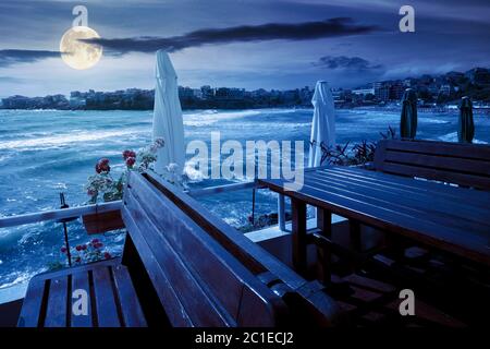 Terrasse eines Restaurants am Meer in der Nacht. Schöne Aussicht in die Bucht im Vollmondlicht. Dekorativer Schirm über Holztischen und Sitzplätzen Stockfoto