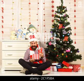 Santa Claus mit Erregten Gesicht in der Nähe von Büro mit Spielzeug und Weihnachtsbaum für den Hintergrund. Feier und lassen Sie sich überraschen. Mann mit Bart öffnet vorhanden. Kerl in Hut und Schal sitzt von Tanne Stockfoto