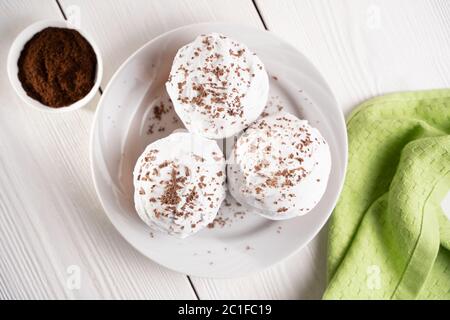 Cupcakes mit Schlagsahne und Schokoladenstückchen auf einem weißen Teller und eine Tasse Kaffee im Hintergrund auf einem weißen Holztisch. Bild für das Menü oder Stockfoto