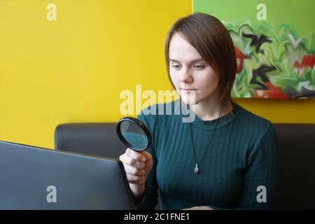 Ein junges, schönes Mädchen mit europäischem Auftritt in einer blauen Jacke, das in einem Café sitzt und auf einem Laptop auf der Couch vor dem Hintergrund einer gelben Wand arbeitet. Betrachtet ein Computerdisplay durch eine Lupe. Layout für Design und Ort für Text. Stockfoto