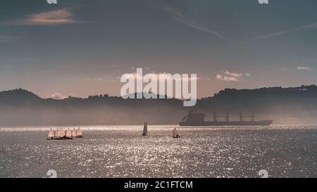 Neblige Seeslandschaft des Flusses Tejo in Lissabon, Portugal mit mehreren Segelbooten während einer Regatta-Sportwettkampf oder einer Performance, mit einem riesigen Schiff Stockfoto