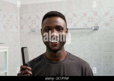 Junger schwarzer Mann lächelt an der Kamera und hält Kamm in der Heimat Badezimmer. Stockfoto