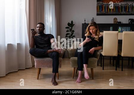 Junge gemischte Rennen Paar im Wohnzimmer sitzen Arme Stühle mit Smartphones weg suchen. Volle Länge. Stockfoto