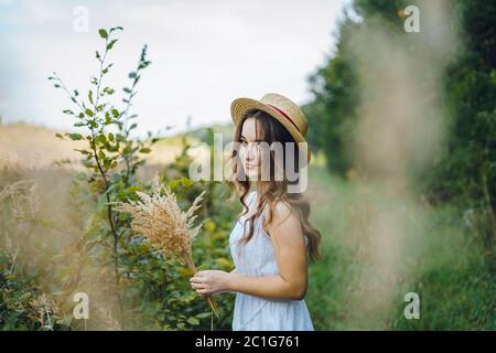 Lächelndes Mädchen in einem Strohhut sammelt Blumen. Mädchen in einem Sommerkleid im hohen Gras. Frau in einem grünen Feld mit einem Bouquet von getrockneten Blumen Stockfoto