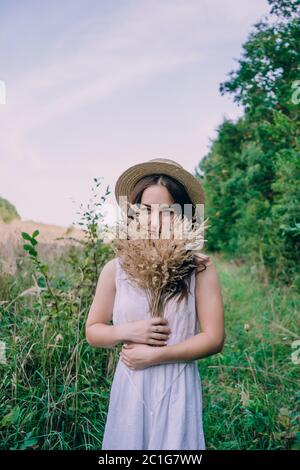 Glückliche junge Frau in einem weißen Kleid in einem Strohhut. Lächelndes Mädchen mit einem Blumenstrauß. Mädchen geht in einem grünen Park an einem Frühlingstag Stockfoto