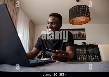 Junger schwarzer Mann lächelt mit toothy Lächeln online line Shopping aus dem Wohnzimmer. Mittlere Aufnahme. Stockfoto