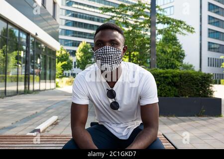 Junger schwarzer Mann mit Gesichtsmaske und Blick auf die Kamera. Mittlere Aufnahme. Im Freien. Stadthintergrund. Stockfoto