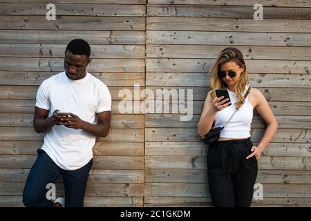 Junger schwarzer Mann und blonde kaukasische Frau mit coolen Einstellungen Blick auf Telefonbildschirme im Freien. Mittlere Aufnahme. Stockfoto