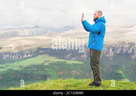 Ein bärtiger Mann in einer membranblauen Jacke steht in der Natur vor der Kulisse der Berge und fotografiert auf dem Tel Stockfoto