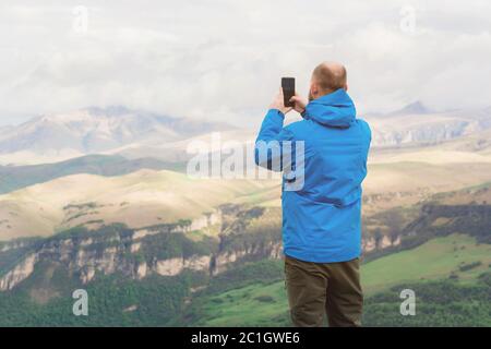 Ein bärtiger Mann in einer membranblauen Jacke steht in der Natur vor der Kulisse der Berge und fotografiert auf dem Tel Stockfoto