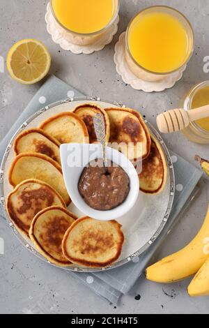 Hausgemachtes Frühstück: Pfannkuchen mit Schokoladenmarmelade, Honig, Bananen und Orangensaft auf grauer Serviette auf betontem Hintergrund, Draufsicht, vertikales Format Stockfoto