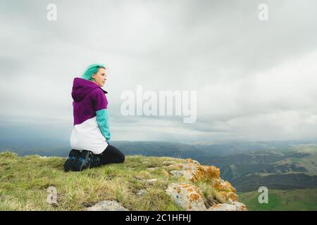 Ein Mädchen-Reisende mit bunten Haaren sitzt am Rande einer Klippe und blickt auf den Horizont auf einem Hintergrund eines felsigen Plateau Stockfoto