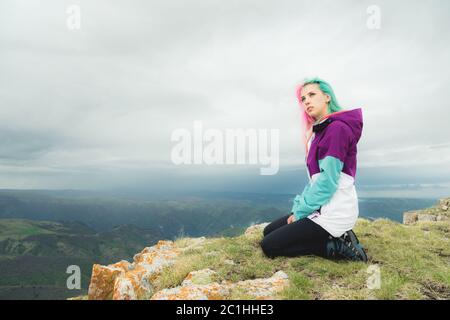 Ein Mädchen-Reisende mit bunten Haaren sitzt am Rande einer Klippe und blickt auf den Horizont auf einem Hintergrund eines felsigen Plateau Stockfoto