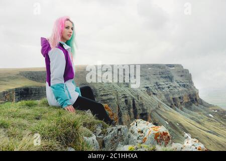 Ein Mädchen-Reisende mit bunten Haaren sitzt am Rande einer Klippe und blickt auf den Horizont auf einem Hintergrund eines felsigen Plateau Stockfoto