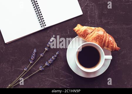 Tasse Kaffee mit Croissant, Notizblock und Lavendelblüten auf schwarzem Hintergrund, Draufsicht, flach liegend Stockfoto