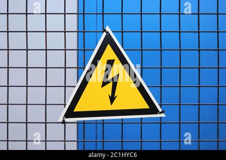 Hochspannungs-Dreieck Warnschild auf einem Metallgitter Zaun auf einem grauen und blauen Hintergrund montiert. Stockfoto