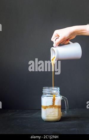 Kaffeeschaum wird in einen Becher mit Milch gegossen. Dalgona Kaffee zubereiten. Schwarzer Hintergrund, Nahaufnahme Stockfoto
