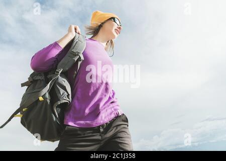 Das Mädchen der Touristen in Sonnenbrille setzt auf einen Rucksack auf dem Hintergrund des Abendhimmels. Vorbereitung für Trekking mit Klettern. Stockfoto