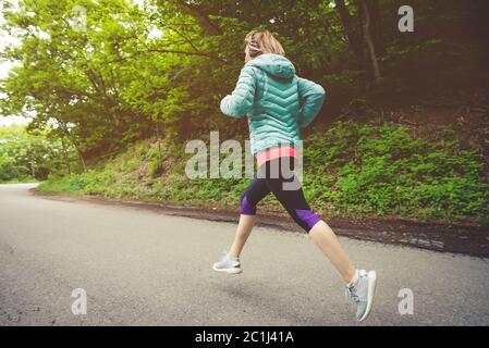 Junge Fitness blonde Frau in Kopfhörer läuft am Morgen kaukasischen Waldweg in Sonnenlicht. Seitenansicht von hinten Stockfoto