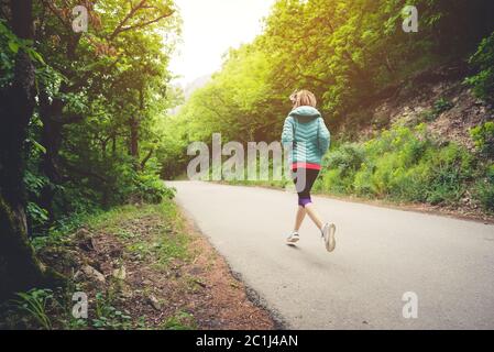 Junge Fitness blonde Frau in Kopfhörer läuft am Morgen kaukasischen Waldweg in Sonnenlicht. Seitenansicht von hinten Stockfoto