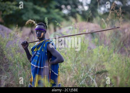 Ein junger Mann aus dem Mursi Stamm im Omo Tal, Äthiopien, trägt einheimische Kleidung und trägt einen Stock, der zum Bewachen von Tieren verwendet wird, und schaut auf die Kamera. Stockfoto