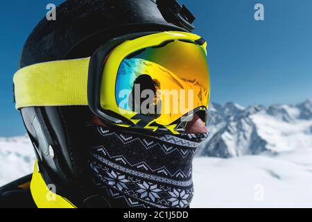 Nahaufnahme eines Skifahrers in Maske und Helm mit geschlossenem Gesicht vor dem Hintergrund schneebedeckter Berge und blauem Himmel Stockfoto