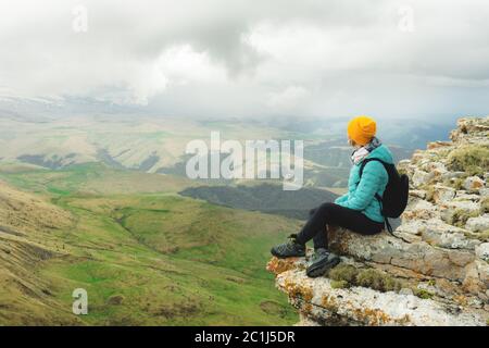 Junge Frau mit Rucksack, die pensiver am Rande eines Felsens sitzt und mit Wolken in den Himmel blickt Stockfoto