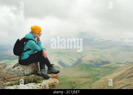 Junge Frau mit Rucksack, die pensiver am Rande eines Felsens sitzt und mit Wolken in den Himmel blickt Stockfoto