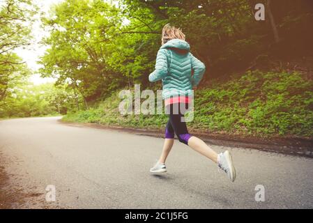 Junge Fitness blonde Frau in Kopfhörer läuft am Morgen kaukasischen Waldweg in Sonnenlicht. Seitenansicht von hinten Stockfoto