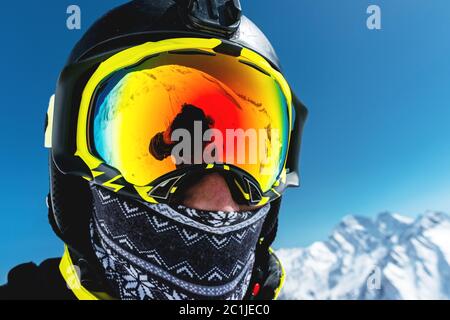 Nahaufnahme eines Skifahrers in Maske und Helm mit geschlossenem Gesicht vor dem Hintergrund schneebedeckter Berge und blauem Himmel Stockfoto