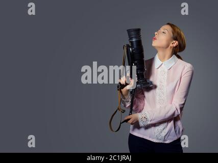 Seitenprofil einer jungen Frau mit einer professionellen Kamera, die sie als Waffe hält und Luft in sie bläst wie in einer Waffe. Gemischtes Rennmodell isoliert auf grauem Rücken Stockfoto